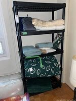 Front angle view of black five-level plastic shelf unit loaded with various folded textiles and bedding items (not included in sale). Shelf surfaces have a grid open pattern supported by four vertical cylindrical legs.