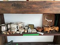 Shelf containing pewter cups, ceramic mugs, ceramic figurines, carved wooden animals, coral stones, and wooden ibex plaque.