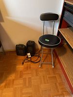 Small black padded stool with backrest and metal legs, two black plug-in electric heaters on floor next to it.