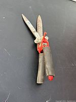Close-up photo of a garden shear with black rubber handles and red locking latch, blades open showing rust and wear.