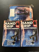 Photo of two boxed hand grip strengtheners with packaging showing brand 'No Excuses' and product description 'Hand Grip Strengthener' with bag included.