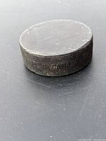 Side close-up showing knurled edge and engraved text 'The Official Game Puck' on a black hockey puck