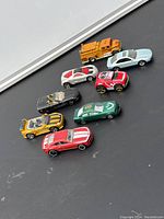 Eight small toy car models in mixed colors and styles on a dark surface.