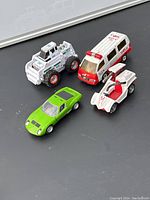 Four toy vehicles on a black surface: green sports car, remnance of a red and white ambulance, white and red racing car, white heavy construction vehicle.