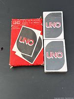 Two sealed decks of UNO cards with classic card backs and red UNO logos, beside the original box.
