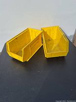 Two yellow plastic hook-on bins with open front on a black surface against a white wall, showing front and side views.