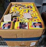 Box full of yellow bordered 1987 Detroit Tigers Coca-Cola baseball trading cards, showing front player portraits on each card, stacked loosely inside a large cardboard box.