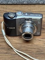 Canon Powershot A1000 IS camera front view, lens extended, with wrist strap on wooden surface.