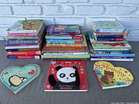 Three shaped board books and three stacks of assorted children's books displayed on floor with a white brick background.