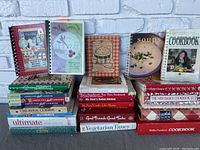 Cookbook lot arranged in stacks on a white brick background showing various titles and colorful covers including Betty Crocker and Better Homes and Gardens.