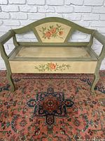 Front view of the painted wooden bench showing floral motifs on backrest and seat lid with curved legs.