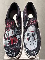 Top-down view of pair of slip-on shoes with Friday The 13th theme, showing distinct graphics on each shoe and paint splatter design.