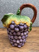 Oblique front view of hand painted porcelain pitcher shaped as grape cluster with leaves and brown handle on a wooden surface with patterned background.