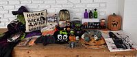 Full view of assorted Halloween decorations on a wooden surface against a white brick wall, including pumpkin figurines, soap bottles, bat decorations, a pumpkin container, a witch pillow, and Halloween-themed bags.
