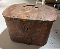 Front and top angled view of a small, old wooden chest with metal straps on the lid and a decorative metal keyhole plate on the front. Wood shows wear, scratches, and some cracking.