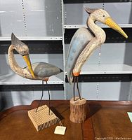 Pair of wooden heron bird sculptures on bases displayed on table with white shelving in background