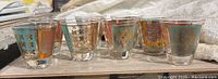 Set of 8 double shot glasses displayed in a row showing colorful sections of teal, red, and gold with intricate coat of arms designs printed in gold foil along the sides.
