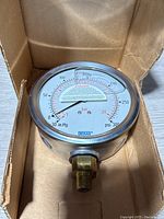 Pressure gauge in original cardboard packaging, showing front dial and threaded brass connector.