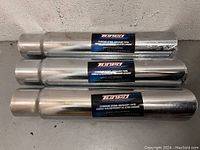Three brand new Tuned chrome steel exhaust pipes stacked, sealed in shrinkwrap with visible labels.
