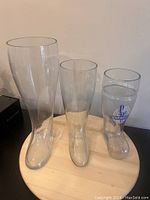 Three glass beer boots of different heights, clear glass, one with logo, shown on wooden surface