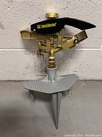 Front view of the Melnor impact sprinkler showing the black and gold metal head with the gray three-prong spike base.