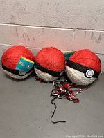 Three Pokéball pinatas sitting on the floor, showing their spherical shape and classic red, white and black color pattern with ribbons attached at the bottom.