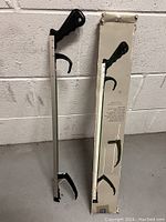 Photo showing the reacher grabber tool standing next to its original box against a white wall with a concrete floor.