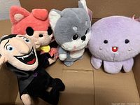 Four stuffed toys in a cardboard box: red fox, gray and white dog, purple octopus, and Dracula doll.