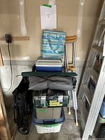 Stacked camping gear items in a garage or basement corner including cooler, foldable chairs, stools, crutches, and storage bins.