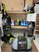 Photo showing Bissell Little Green portable carpet cleaner, Bissell upright vacuum, Hoover Aerovac upright vacuum, and cleaning supplies on shelf.