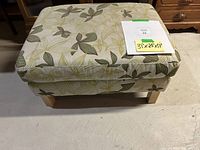 Photo of rectangular IKEA ottoman with light fabric featuring green and beige leaf pattern. Wooden legs visible, piece resting on floor with a note showing measurements 31x24x17 inches.