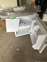 White mini fridge open, showing interior shelving and small freezer compartment, with two door shelves and empty interior, photographed standing on a concrete floor.