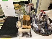 Photo showing various vintage cutlery items on a white surface, including a box of knives in a wooden knife block and some loose cutlery in plastic bags, against a backdrop of dishes and glassware.