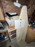 Photo showing a vintage ironing board with floral cover leaning against a surface, a white metal drying rack folded and standing next to it, and a white plastic laundry basket on a small wooden shelf filled with various folded towels.