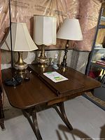 Photo showing three vintage lamps of varying styles placed on top of a wooden vintage drop-leaf table with curved legs, situated in a basement with unfinished walls.