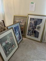 Photo shows six large vintage art prints in frames leaning against a wall, including floral and village scenes.