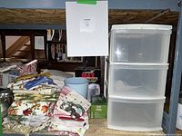 Photo showing a stack of colorful vintage linens next to a white plastic three-drawer storage bin on a wooden shelf in a basement setting.