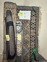 Photo showing three rolled or partially unrolled vintage rugs stacked overlapping on concrete floor, with visible size stickers 6'1 x 3'3, 10 x 10, and 4'8 x 2'4.