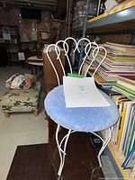 Full view of vintage white metal framed vanity chair with blue round upholstered seat in basement cluttered with books and miscellaneous items.