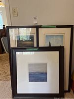 Three framed artworks leaned against wall showing various blue and neutral-toned landscapes and abstract scenes.