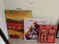Three framed paintings arranged on floor: abstract figures in foreground, motorcycle in middle, landscape in back.