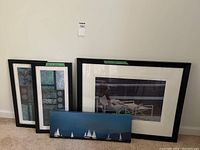 Five framed artworks shown together leaning against a white wall on carpeted floor.