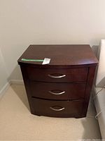Front view of one dark wood bedside table showing three drawers with silver curved handles, used condition.