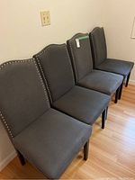 Four grey fabric dining chairs arranged side by side, showing cushioned seating, studded trim on backrests, and dark wooden legs.