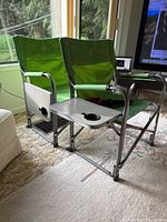 Two green folding chairs with metal frames placed inside near window, showing attached side trays with cup holders.