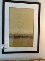 Framed large artwork showing abstract horizontal brown lines on a gold background within a dark wooden frame.