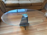 Full view of oval beveled glass coffee table with metal stand positioned on wood flooring in front of a couch.