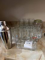 Various clear glassware including highball, tumblers, and shot glasses along with a stainless steel martini shaker and rectangular glass butter dish with lid displayed on a counter.
