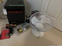 EdenPure Quartz Infrared portable heater, white desk fan, two lanterns, flashlight, and batteries arranged on carpeted floor.