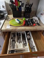 Wide shot showing the countertop with utensils in black holder, two knife blocks, cutting board with small tools, and an open drawer with silverware organizer.